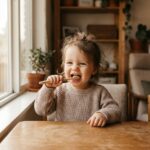 Toddler smiling while holding a toothbrush at a wooden table, illustrating early oral health habits during the first 1,000 days